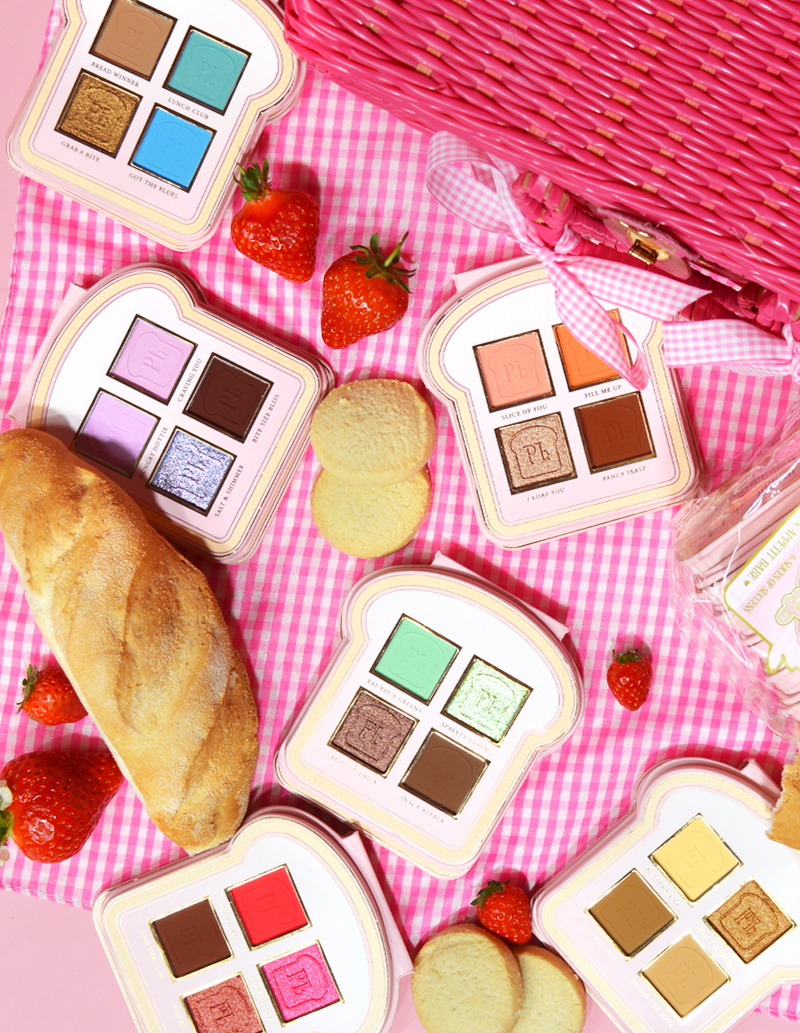 En uppsättning ögonskuggspaletter i mini-sandwich-stil med olika nyanser av sminkfärger som visas på en rosa gingham-yta, tillsammans med jordgubbar och en bit bröd.
