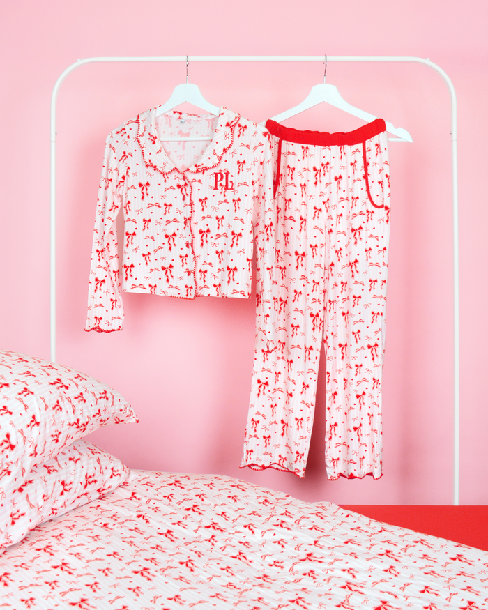 Girls P.Louise Christmas rosett pyjamasbyxor set med en hel röd rosett design mot rosa och vita ränder som hänger på klädhängare i ett rosa sovrum med matchande sängkläder