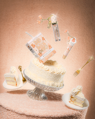 En tårta på ett teddytygsbord med en P.Louise Teddy Glitter Bad Bitch Energy Lip Duo i kokosnötskaka innehållande läppbalsam och läppolja som flyter runt tårtan 