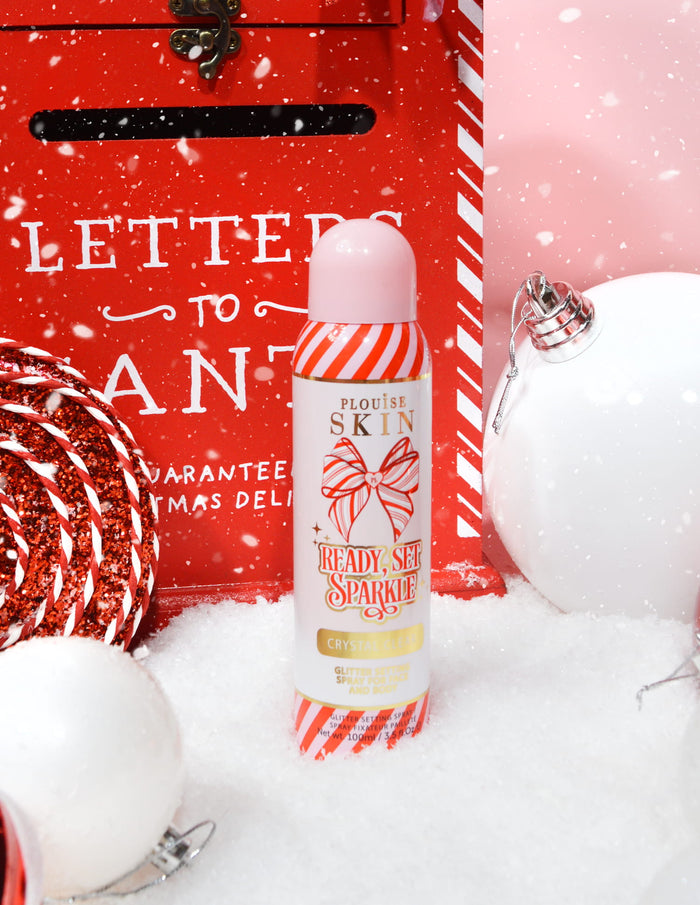 En flaska P. Louise Ready Set Sparkle Christmas Glitter Setting Spray omgiven av festliga dekorationer som snö, grannlåt och en julbrevlåda