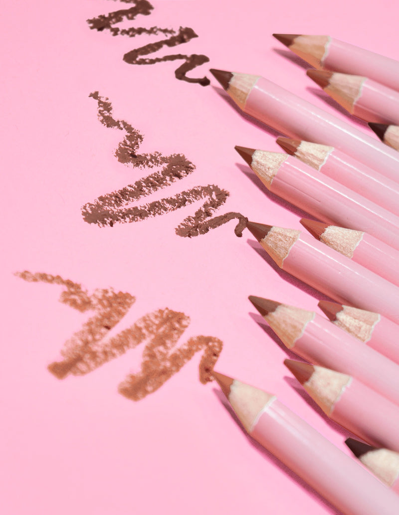 En kollektion av P.Louise ultimate definition slipbara läppliners i en mängd olika nude nyanser, utlagda på en rosa bakgrund med några linjer av produkten ritade på ytan.