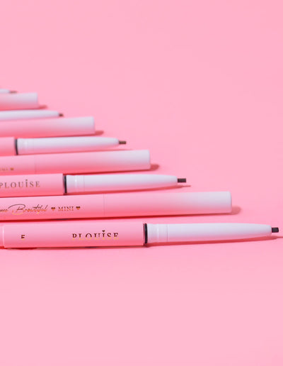 En samling P.Louise Mini Eyebrow Pencils läggs på rad med varumärket synligt på pennornas kropp, visat mot en rosa bakgrund