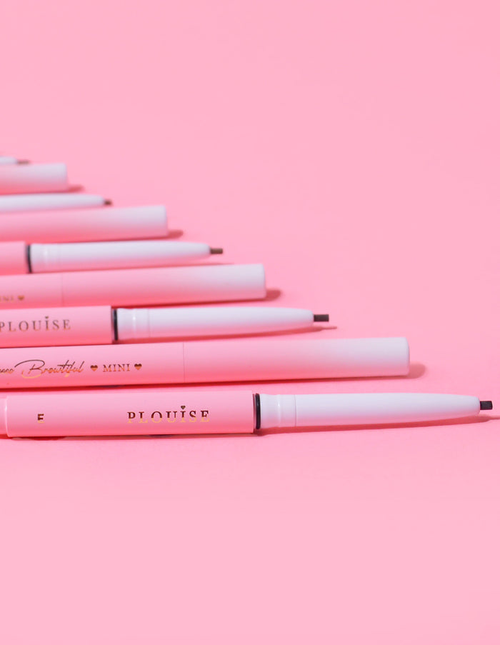 En samling P.Louise Mini Eyebrow Pencils läggs på rad med varumärket synligt på pennornas kropp, visat mot en rosa bakgrund